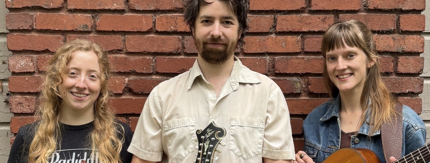 Flat Bridge: A man stands in front of a red brick wall holding a mandolin. Two women are beside him; one is holding a fiddle, the other an acoustic guitar.